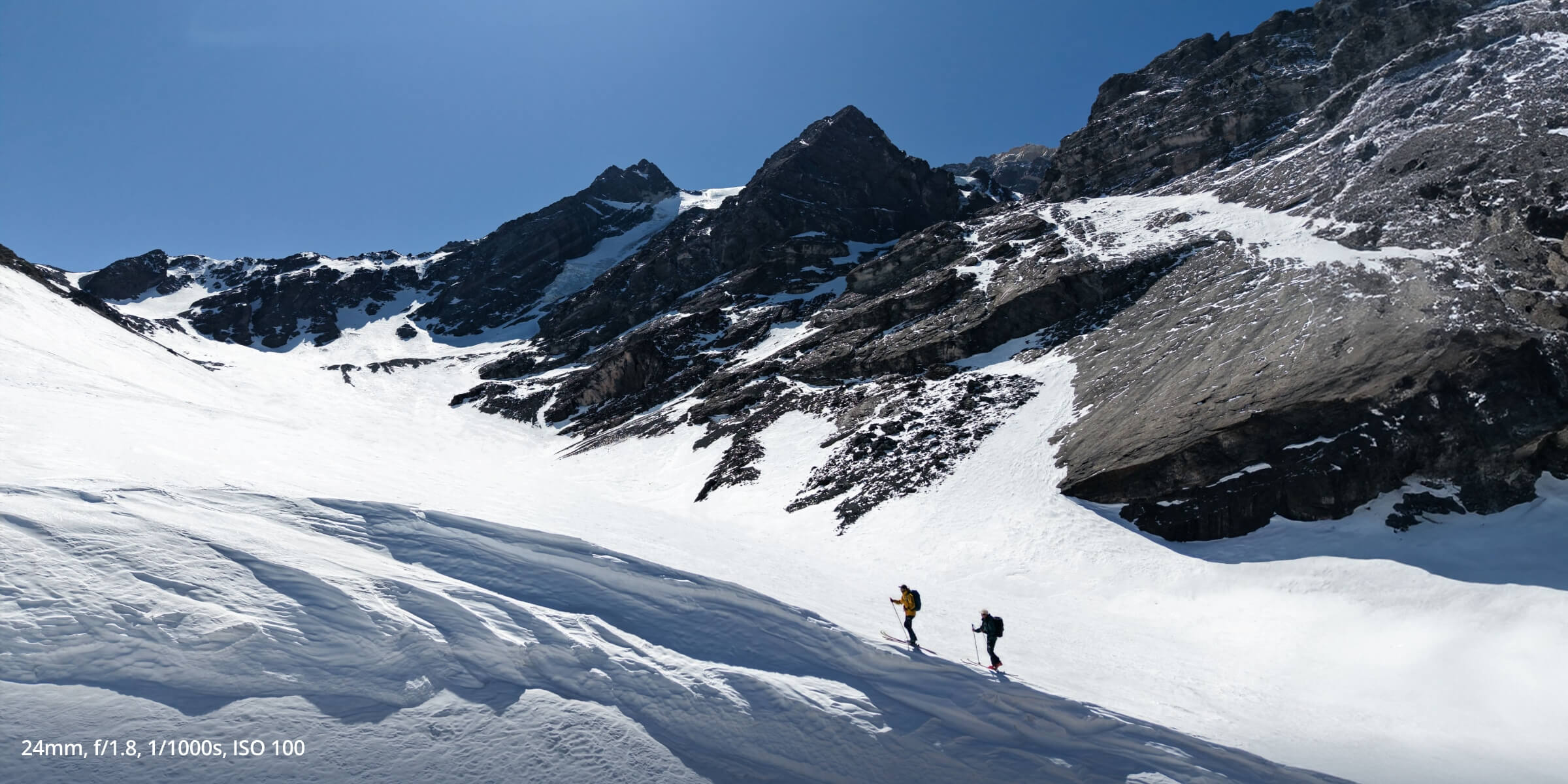 Air 3S photo of 2 hikers walking up snow covered mountains, taken with 24mm, f/1.8, 1/1000s, ISO 100
